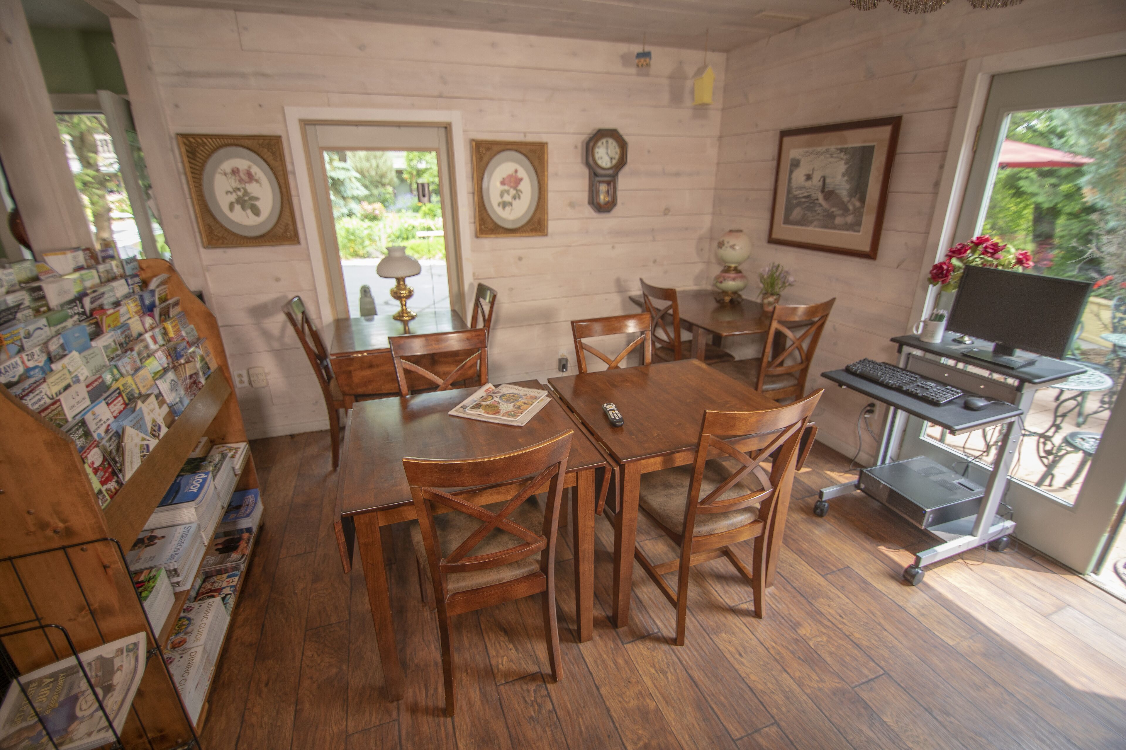 Een eetgedeelte met houten tafels en stoelen, een computer bureau met monitor, toetsenbord en muis, en een wandklok.