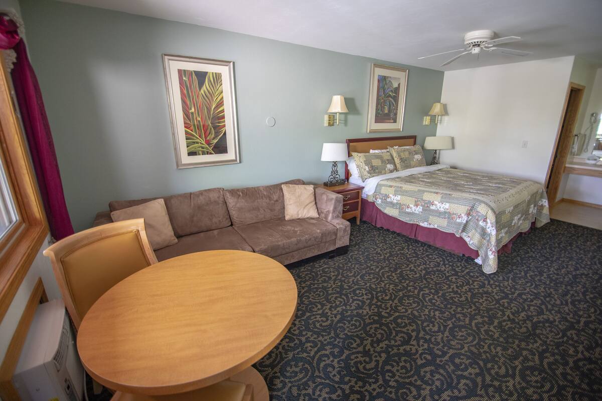 A hotel room with a sofa, a round wooden table, a bed with a patterned bedspread, two wall-mounted lamps, and two framed artworks on the walls.