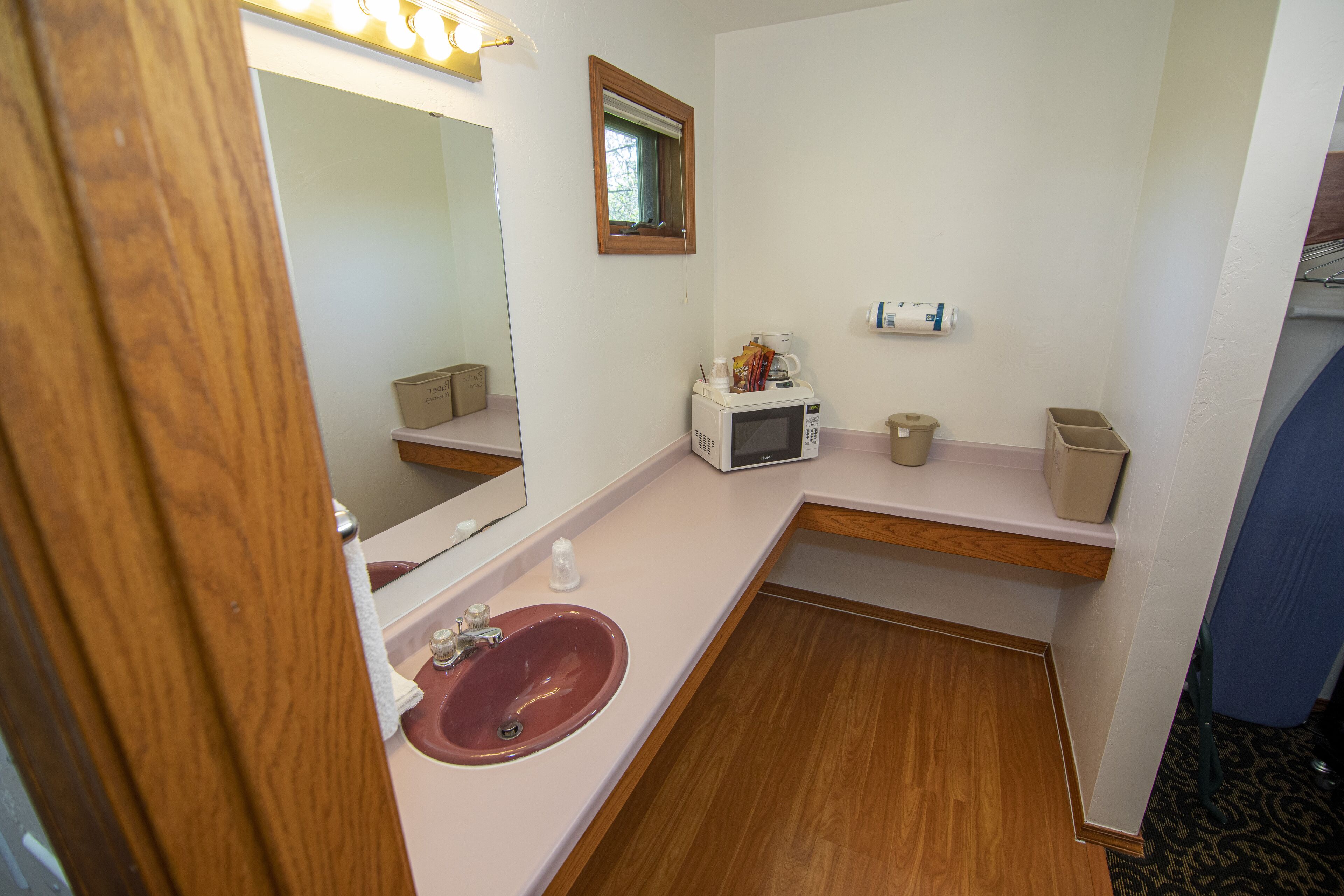 Un baño con tocador de madera, lavamanos, espejo, microondas y un estante pequeño con artículos de aseo.