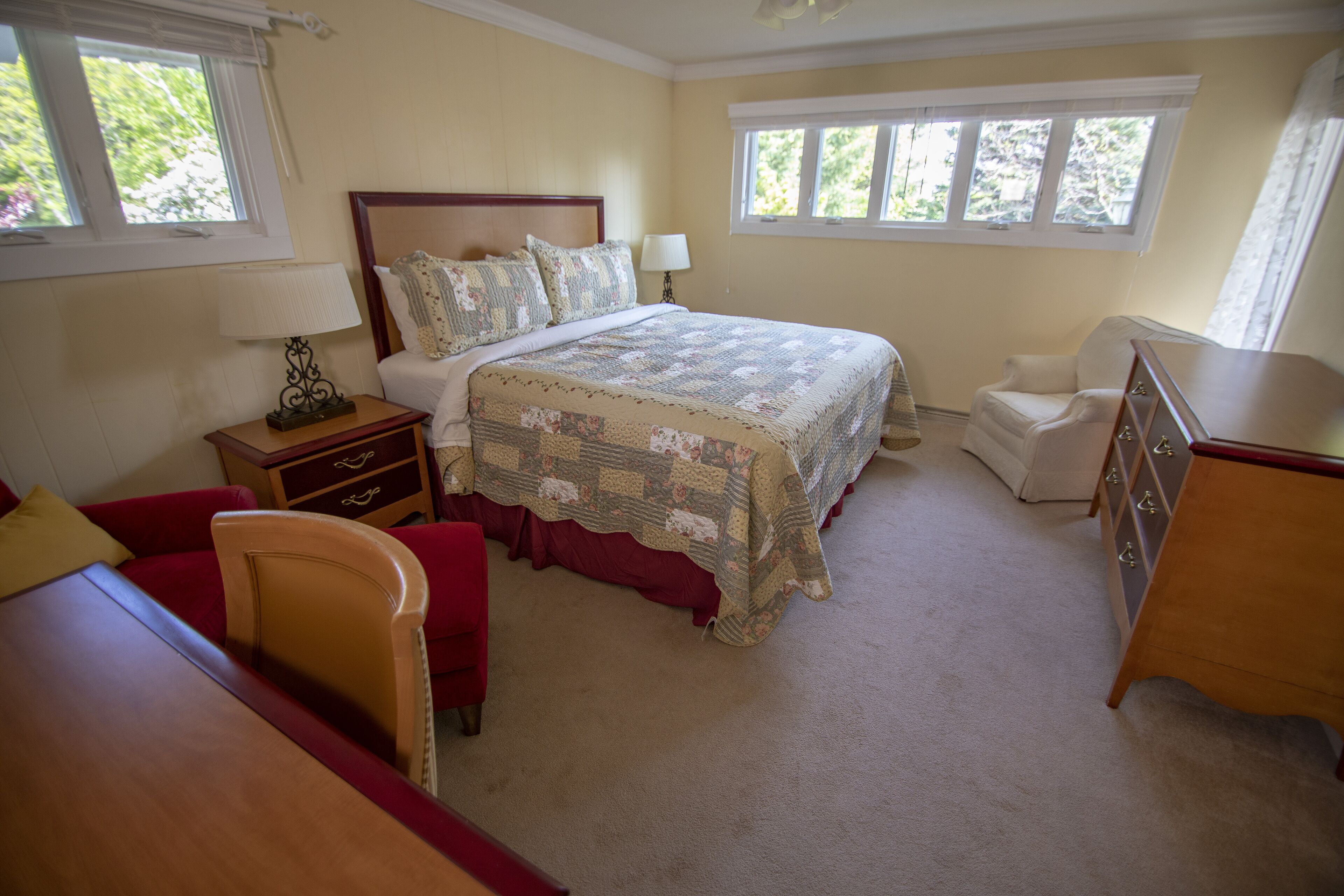 A bedroom with a large bed, a bedside table with a lamp, a chair, a dresser, and a window with a view of greenery.