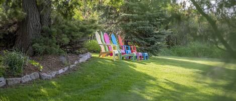 Una hilera de sillas Adirondack de colores en un área verde con árboles al fondo.