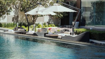 Un área junto a la piscina con sillones de descanso y sombrillas, una piscina cristalina y un edificio moderno al fondo.