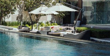 Un área junto a la piscina con sillones de descanso y sombrillas, una piscina cristalina y un edificio moderno al fondo.
