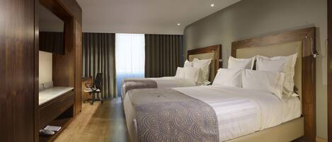A hotel room with two beds, a wooden wardrobe, a desk with a chair, and a large window with curtains.