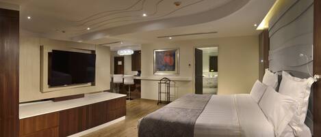 Una habitación de hotel moderna con un televisor de pantalla plana grande, una cama con sábanas blancas, un escritorio con silla y un baño visible a través de una puerta abierta.