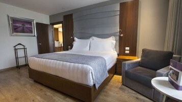 Quarto de hotel moderno com uma cama grande, uma poltrona cinza estofada, uma pequena mesa e um quadro na parede.
