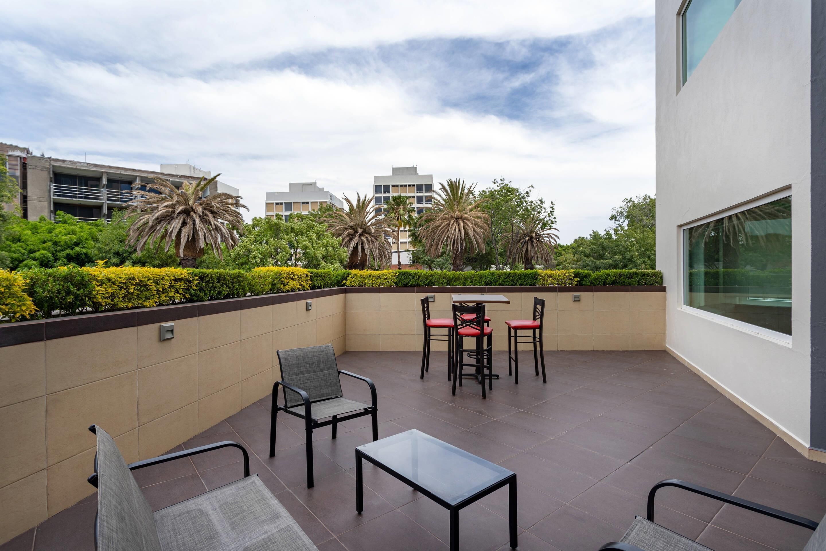 An outdoor patio area with a table and chairs, surrounded by greenery and modern buildings in the background.