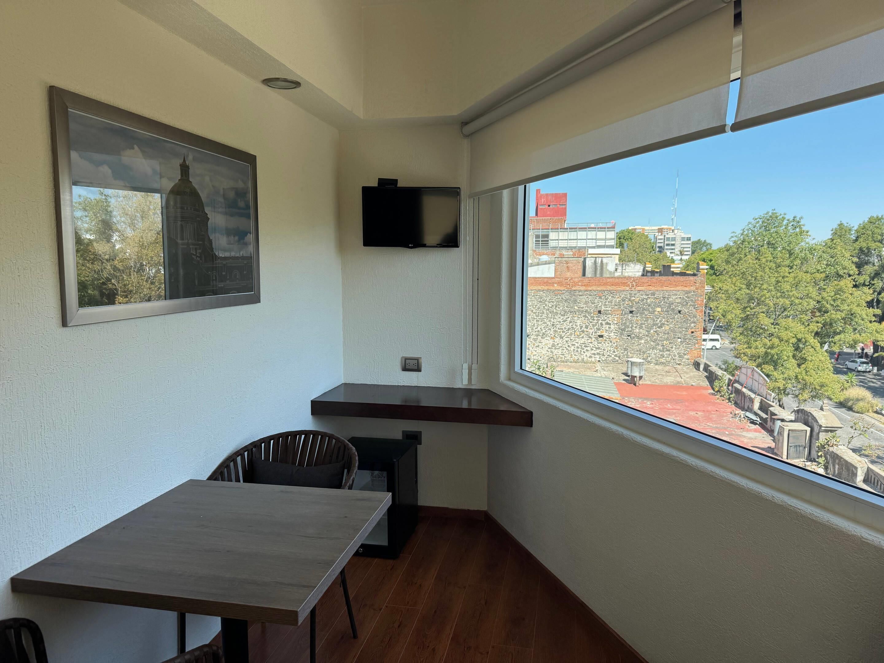 A small room with a wooden table, a chair, a TV mounted on the wall, and a large window offering a view of buildings and trees.