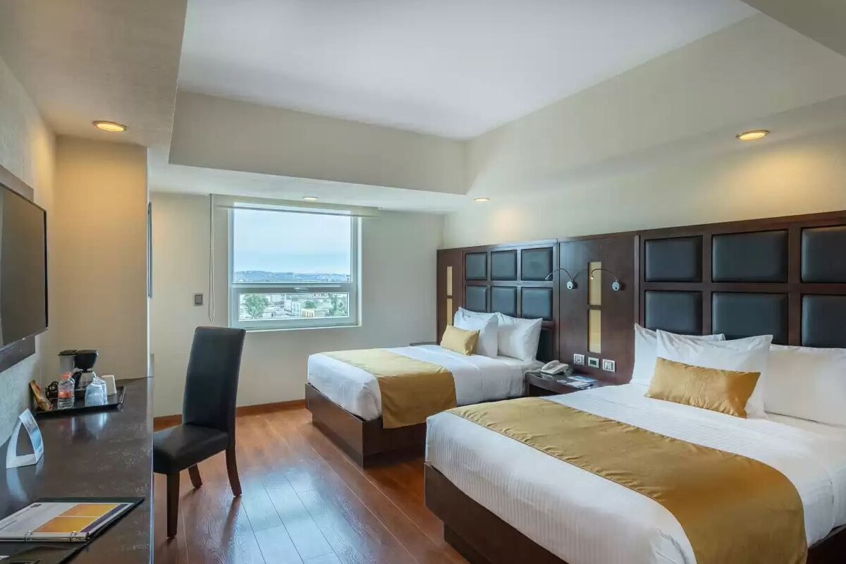 A hotel room with two beds, a desk, a chair, and a window with a view.