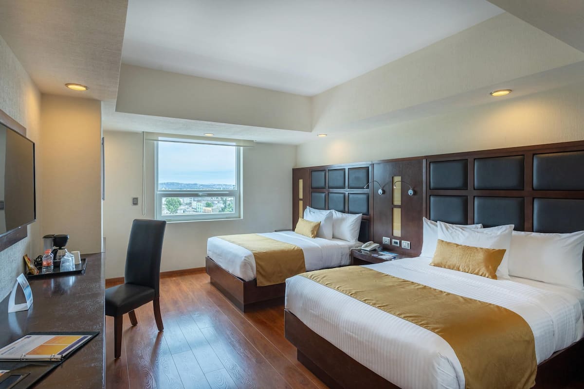 Habitación de hotel con dos camas, un escritorio, una silla, un ventanal con vista a la ciudad y un cabecero de madera.