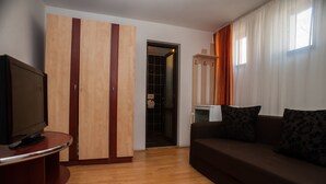 A modern living room with a wooden floor, a brown sofa with patterned cushions, a TV on a curved stand, a wardrobe with vertical stripes, and a window with curtains. - Ceramica Hotel (Iasi)