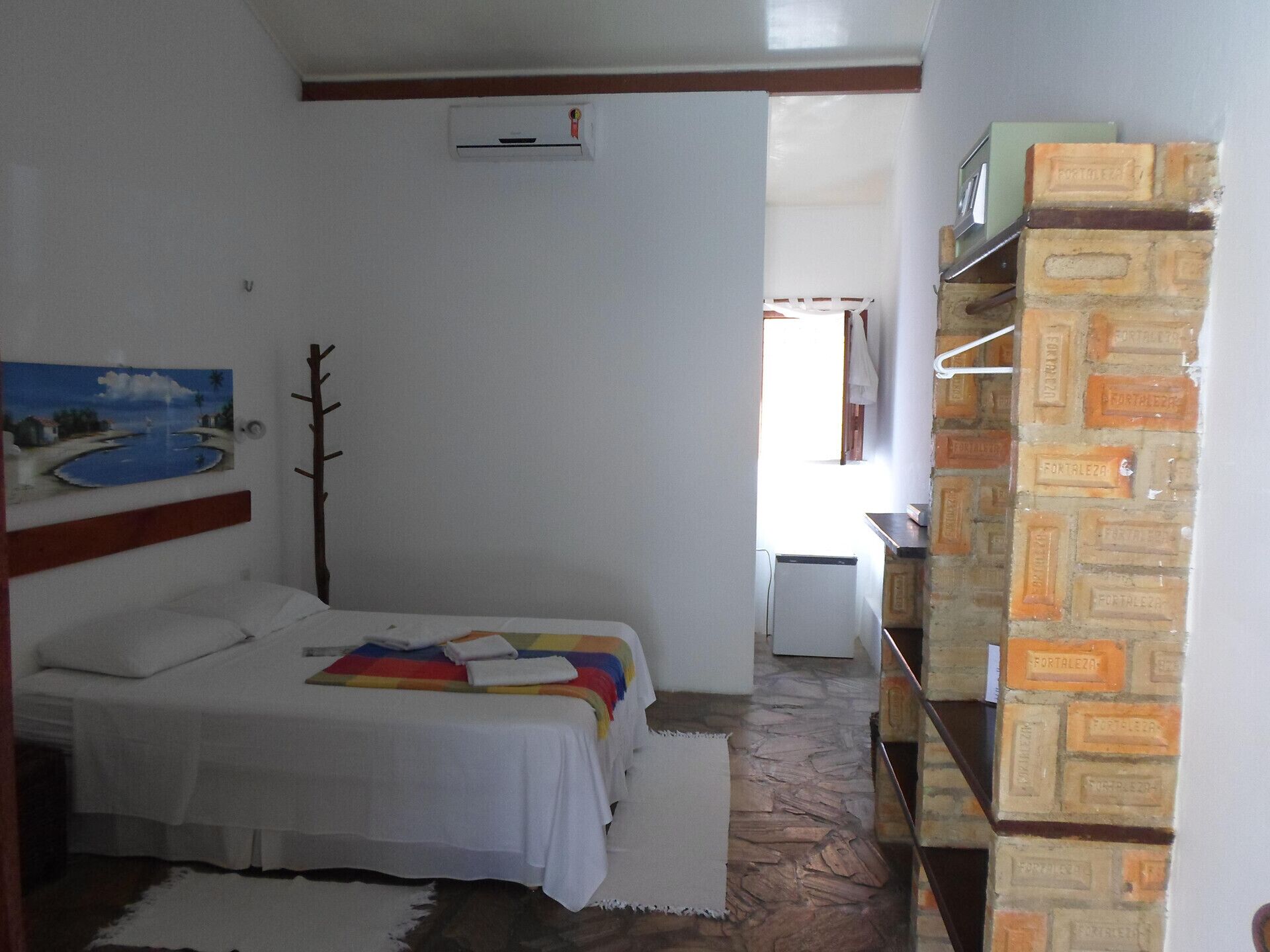 A bedroom with a bed, a painting, a wooden headboard, a white wall, a brick wall, and a window.
