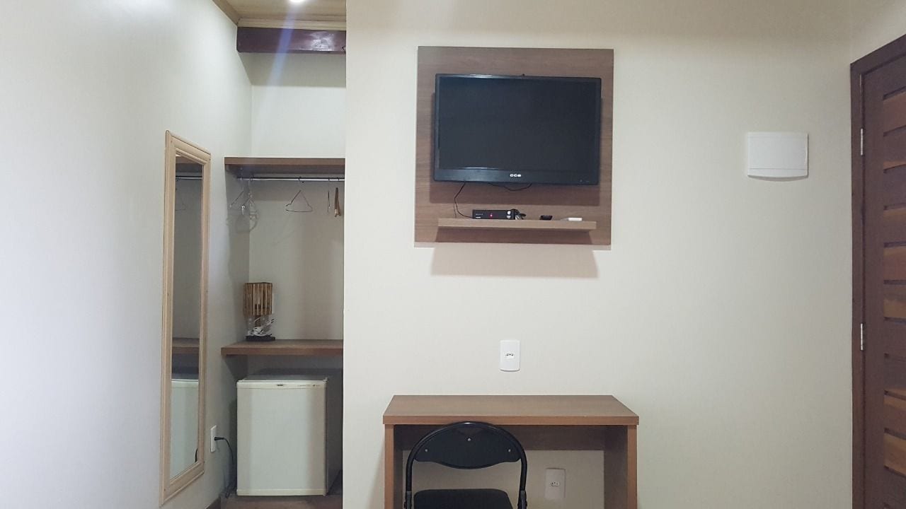 A compact room with a TV mounted on the wall, a small desk with a chair, a mini-fridge, and a wardrobe with a mirror.