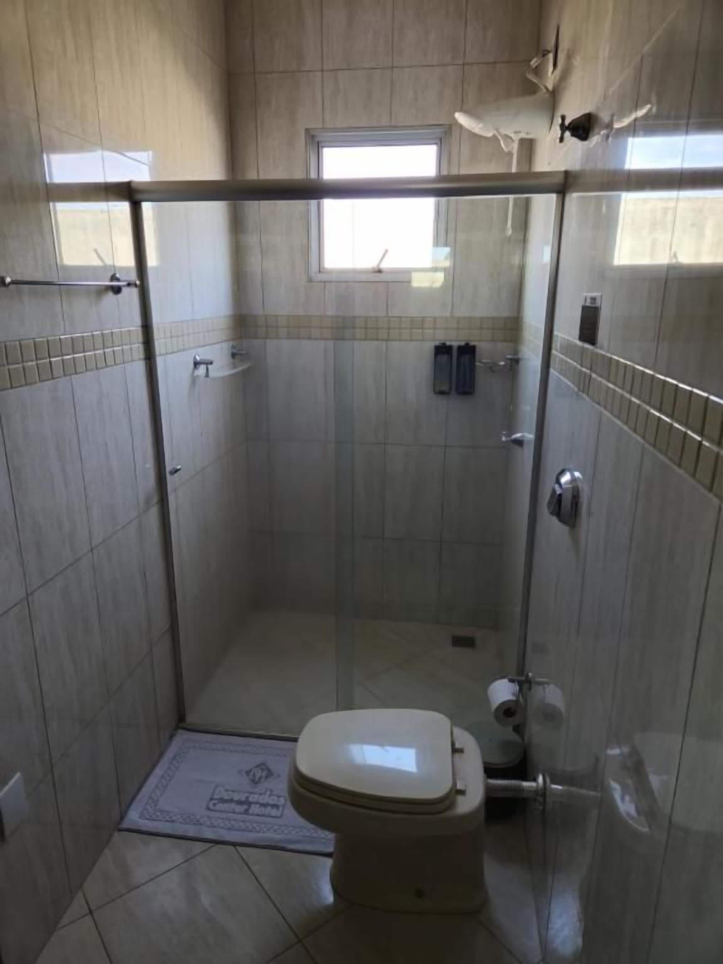 Pequeno box de chuveiro fechado, com vaso sanitário, porta-toalhas e janela que permite entrada de luz natural.