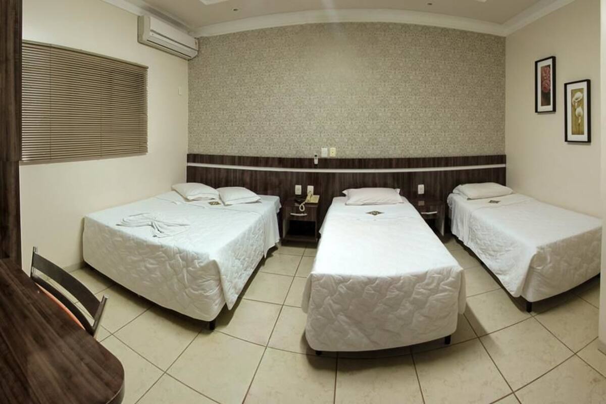 Quarto de hotel com três camas de solteiro, uma mesa de jantar de madeira e uma pequena escrivaninha com telefone.