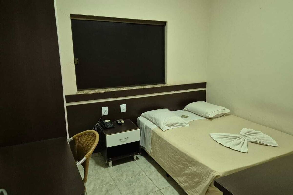 Quarto pequeno e limpo, com uma cama, uma cadeira, uma mesa e uma janela.