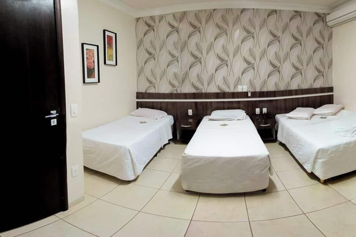 Quarto de hotel com três camas de solteiro, cabeceira de madeira, piso ladrilhado e uma pequena mesa.