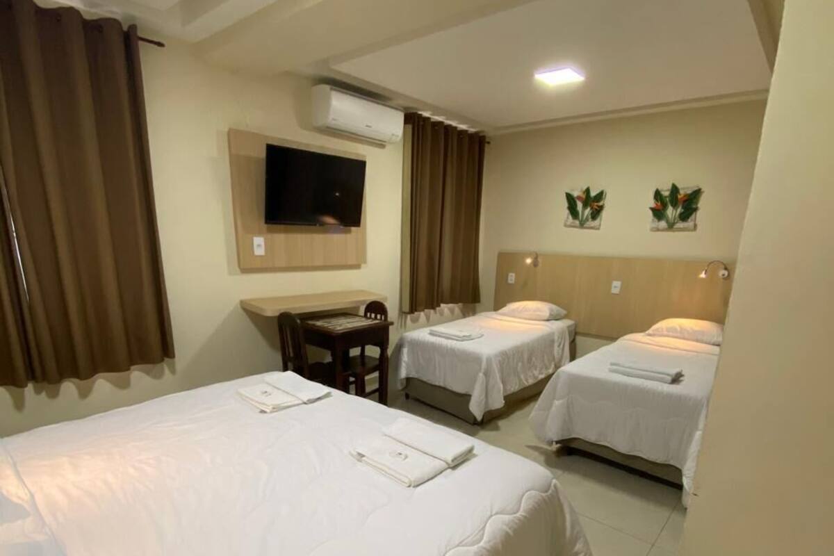 A hotel room with a large bed, two smaller beds, a desk with a chair, a flat-screen TV, and an air conditioning unit.