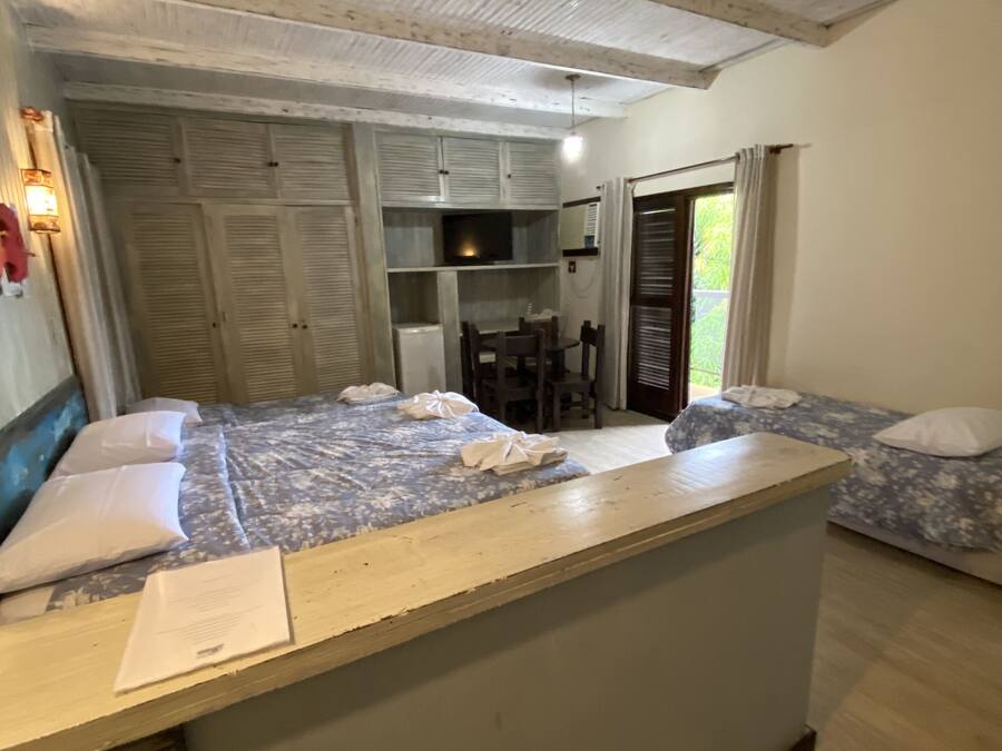 A room with a wooden ceiling, a bed with a blue floral bedspread, a small dining table with chairs, and a kitchenette area with cabinets and a stove.
