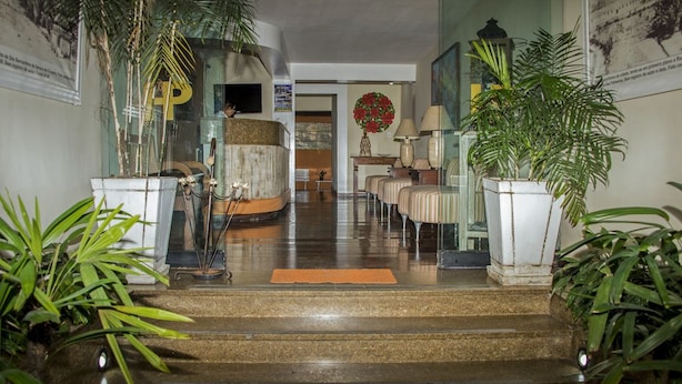 A hotel lobby with a staircase leading to the upper floor, potted plants, a reception desk, and a wall with a large poster. - Palace Hotel Angra (Angra dos Reis)