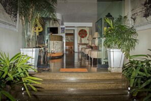 A hotel lobby with a staircase leading to the upper floor, potted plants, a reception desk, and a wall with a large poster.