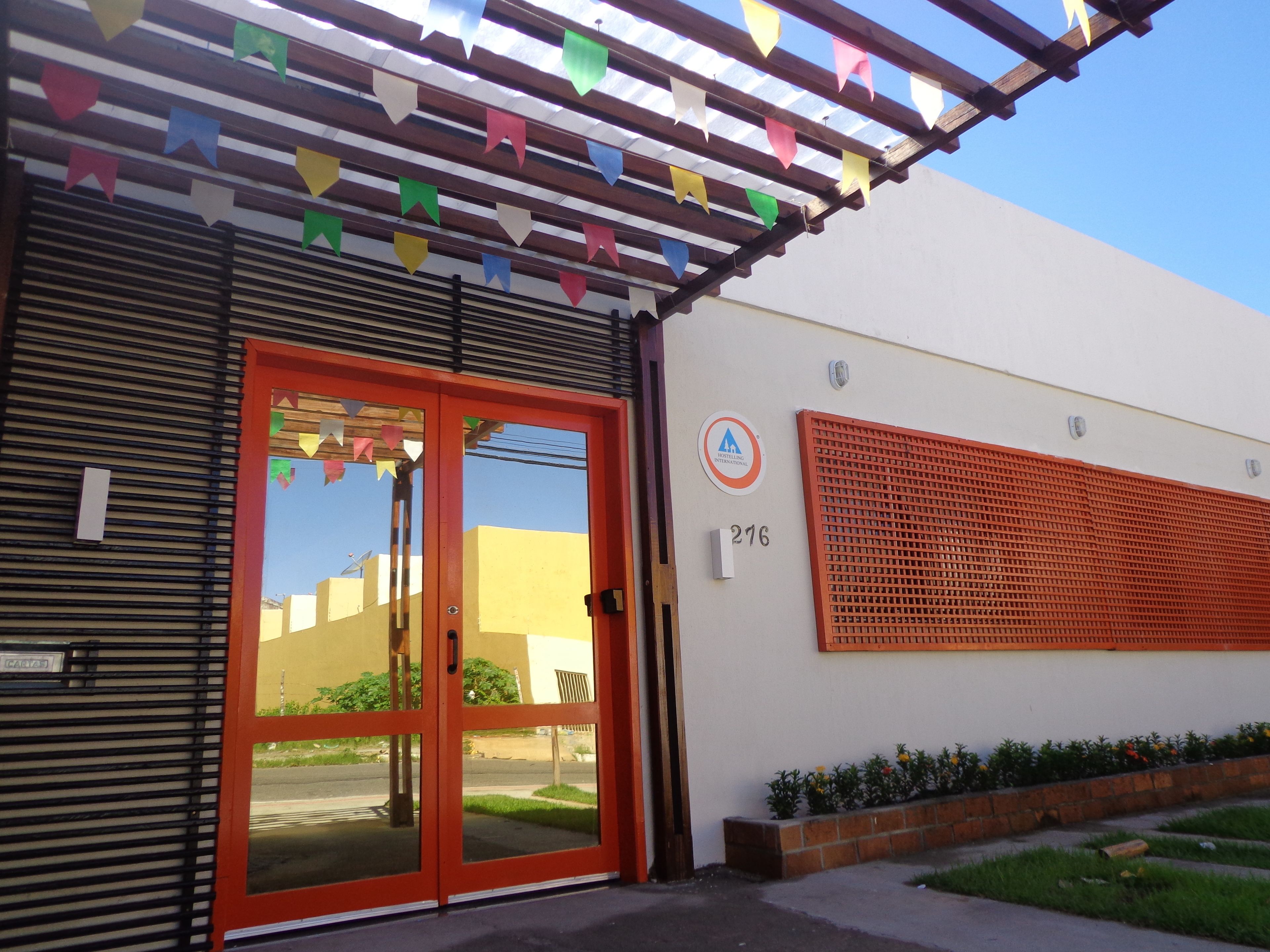 Entrada de un edificio con una puerta roja, un cartel con el número 276 y banderas de colores arriba.