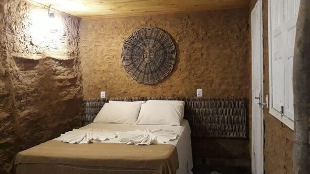 A rustic room with a stone wall, a woven wall hanging, a bed with white linens, and a wooden ceiling. - Vila Aju Pousada Temática (Aracaju)
