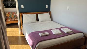 En snyggt bäddad säng med ett lila överkast och två hopvikta handdukar, en sänggavel i trä och en utsikt över en färgglad soffa och en tegelvägg i bakgrunden.