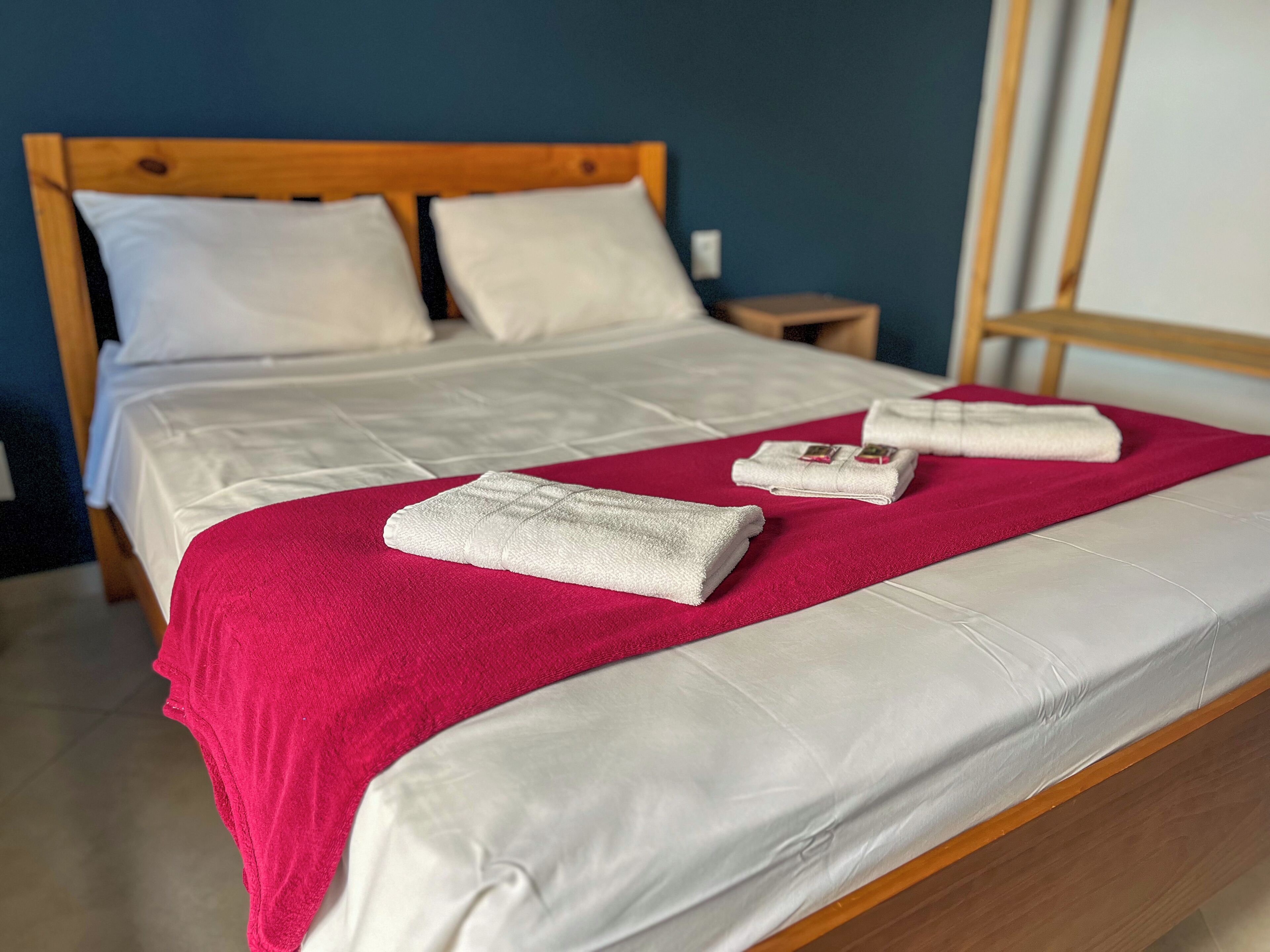 Una cama bien hecha con sábanas blancas y una manta roja, junto con toallas dobladas.