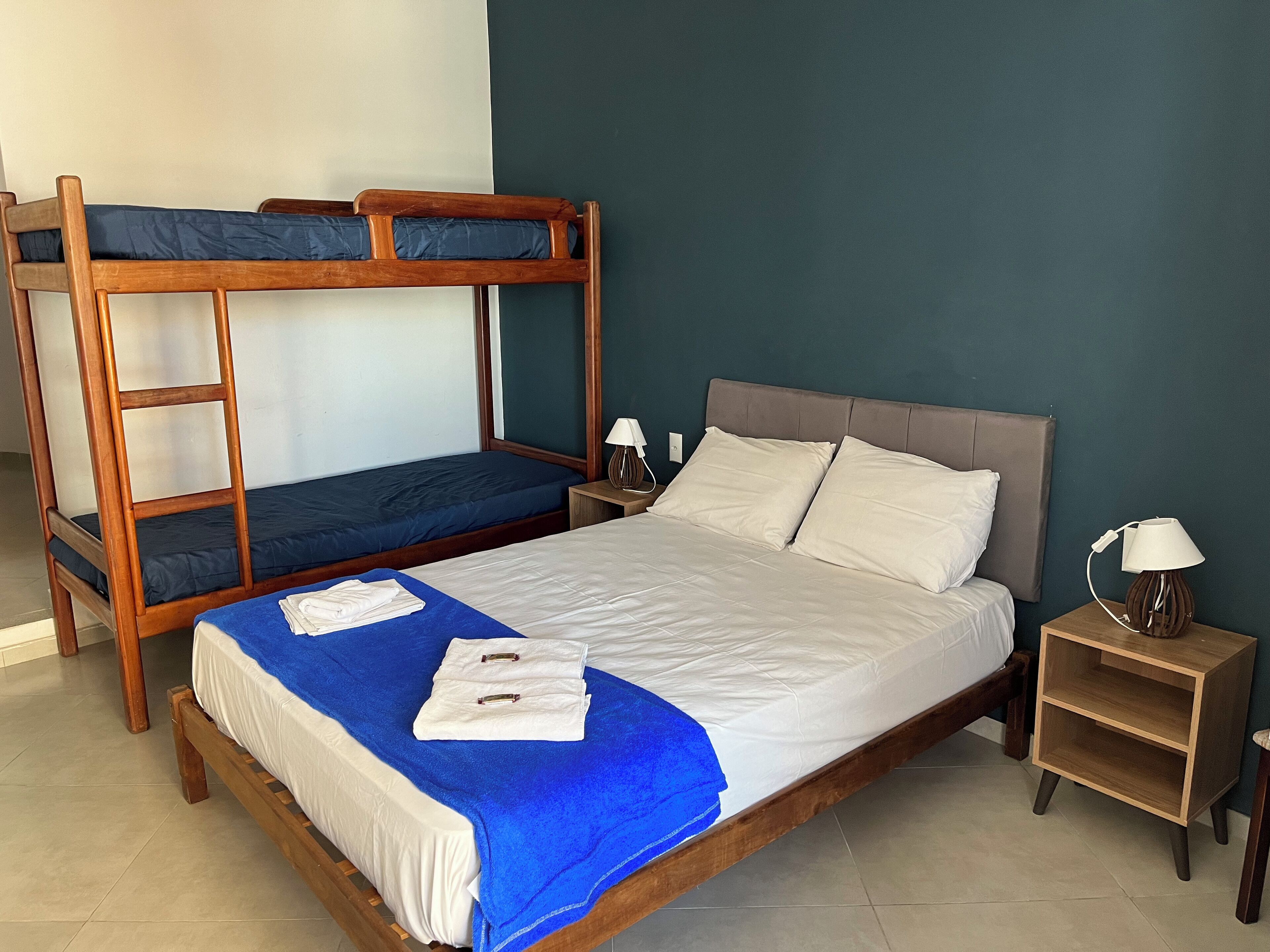 A room with a bunk bed, a single bed, a nightstand with a lamp, and a small wooden shelf.
