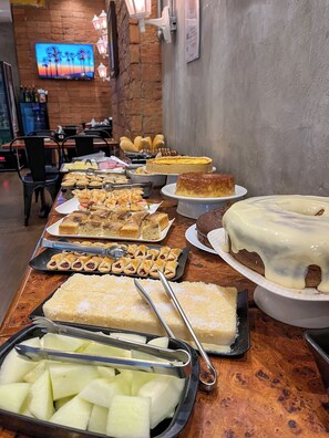 A buffet table with various desserts and a TV screen displaying a sunset scene. - Pousada Vila Appia (Cabo Frio)