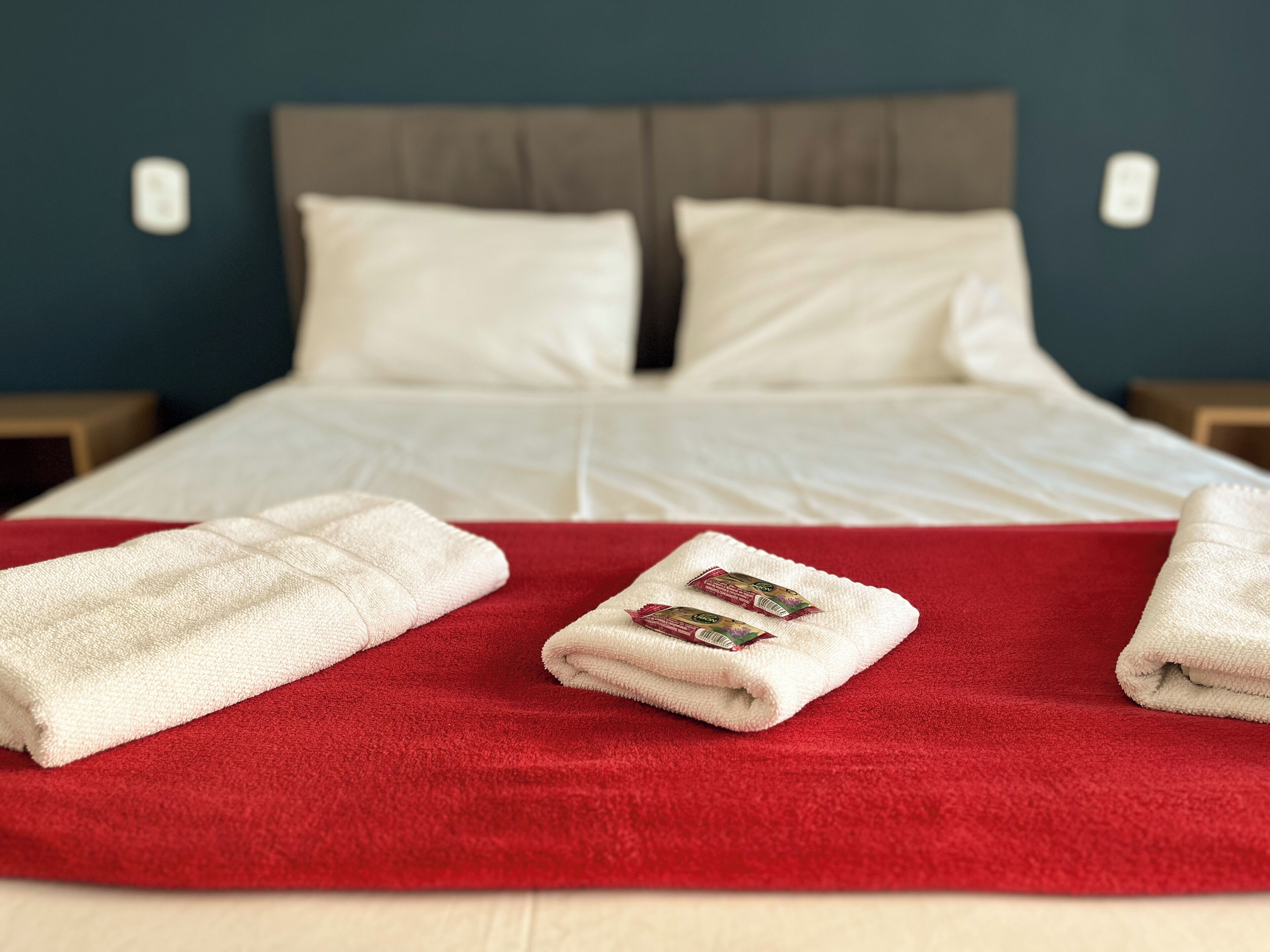 A neatly made bed with white linens, a red blanket, and two folded towels on a red pillow.