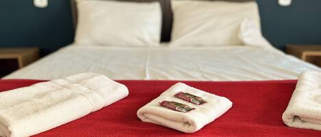 A neatly made bed with white linens, a red blanket, and two folded towels on a red pillow.