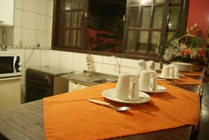 A kitchen with a dining area. There are two white cups on saucers with spoons in front of them. A window with a view of plants is visible in the background. - Pousada Martin Pescador (Bombinhas)