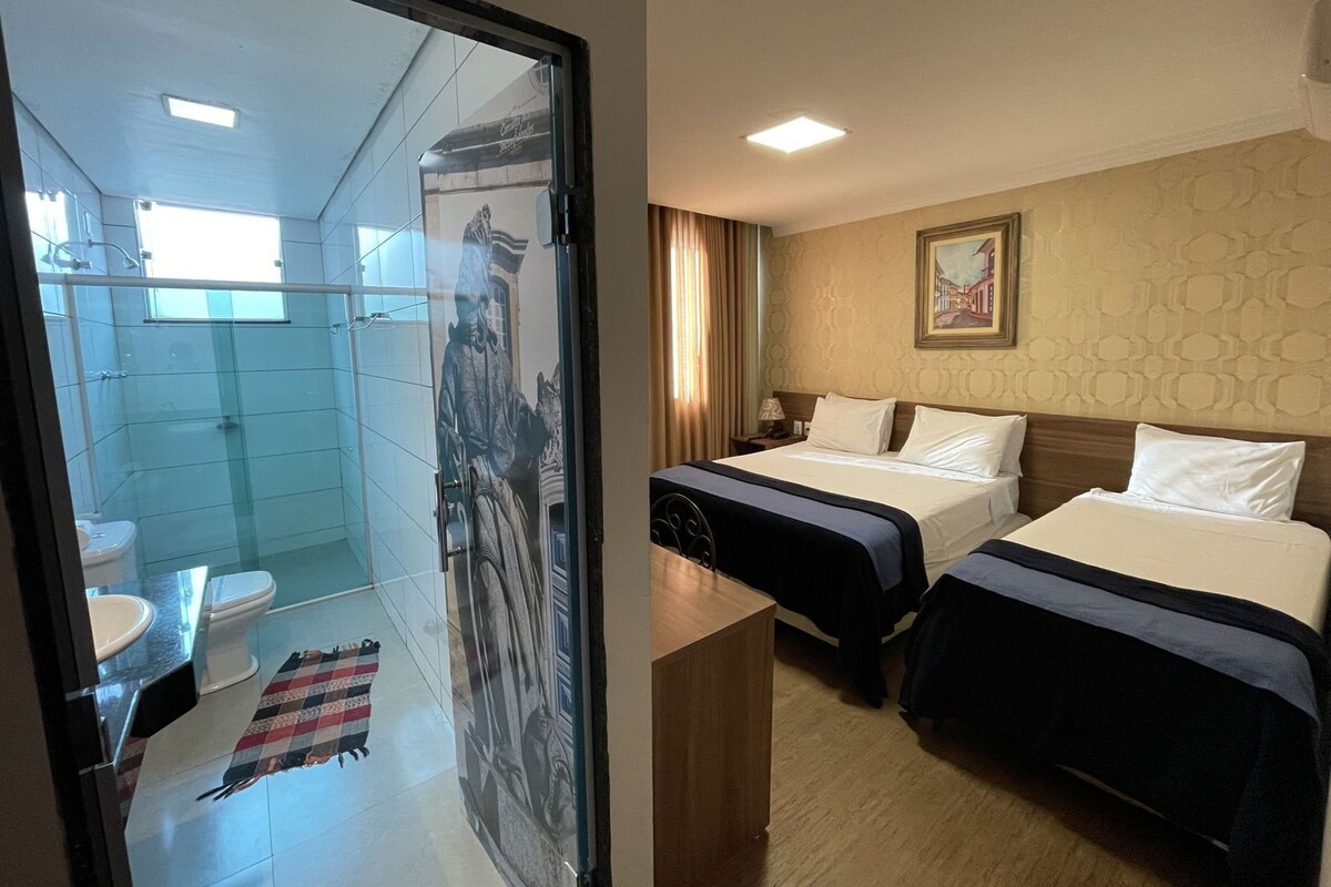 A hotel room with a glass shower enclosure, a bed with two pillows, a nightstand, and a painting on the wall.