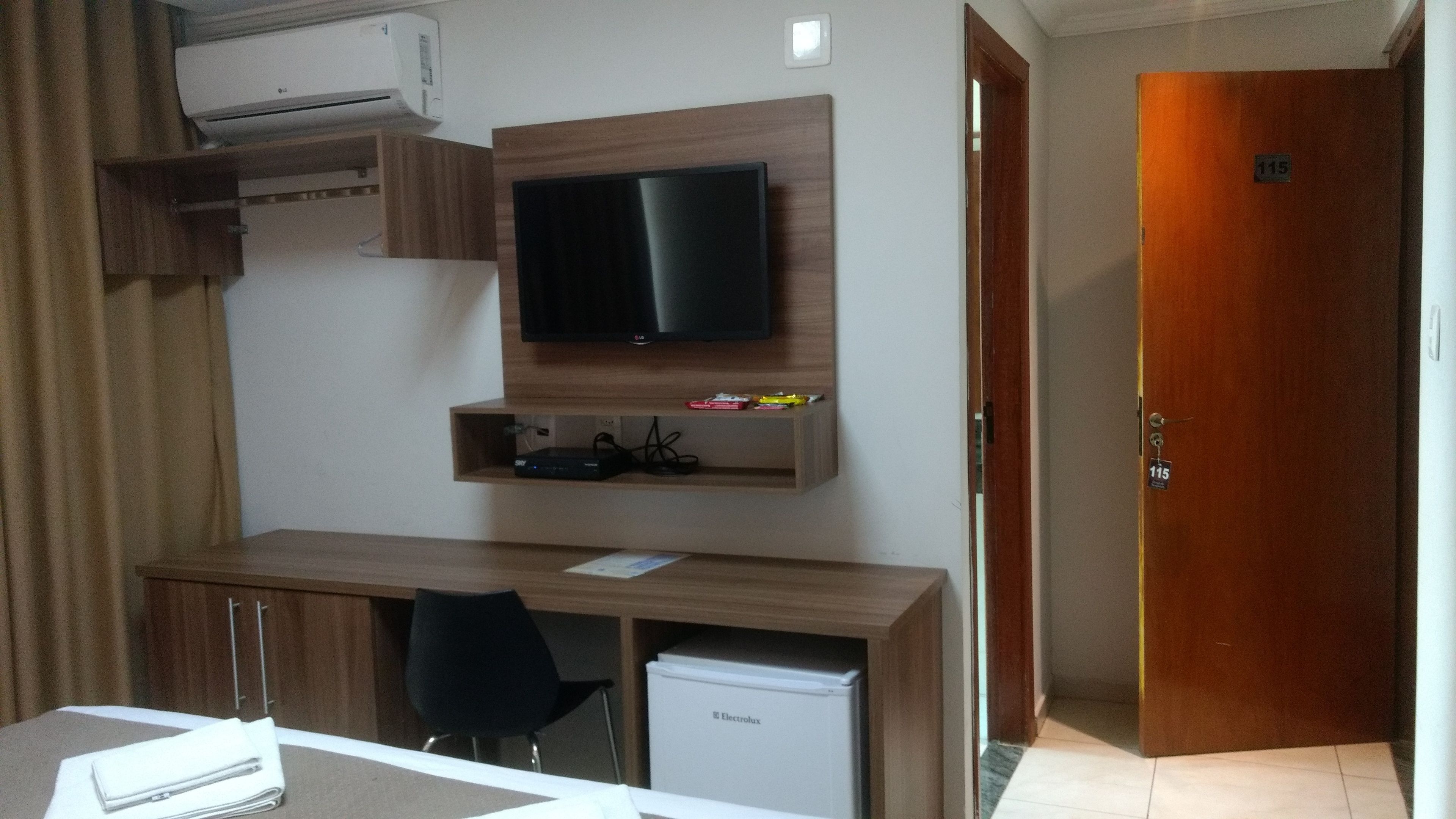 A hotel room with a wooden desk, a television mounted on the wall, a small refrigerator, and a door with a number.