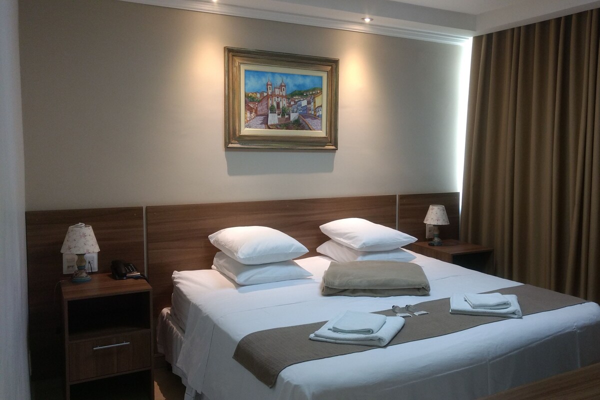 A hotel room with a bed, pillows, a nightstand, a lamp, a painting on the wall, and a window with curtains.