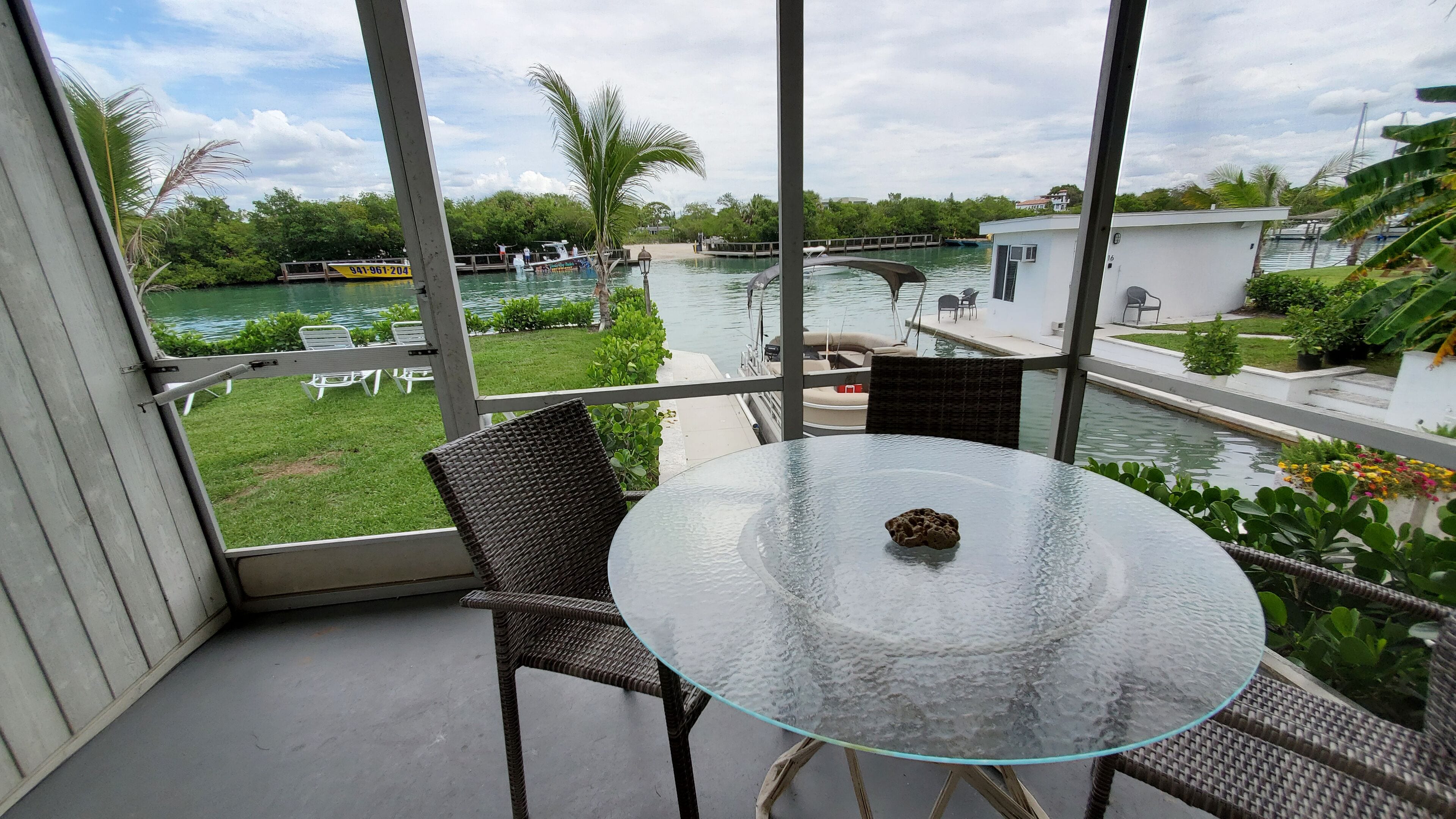 Une table à plateau en verre, accompagnée d’une chaise en osier, sur un balcon donnant sur un plan d’eau où l’on peut voir des bateaux et de la végétation.