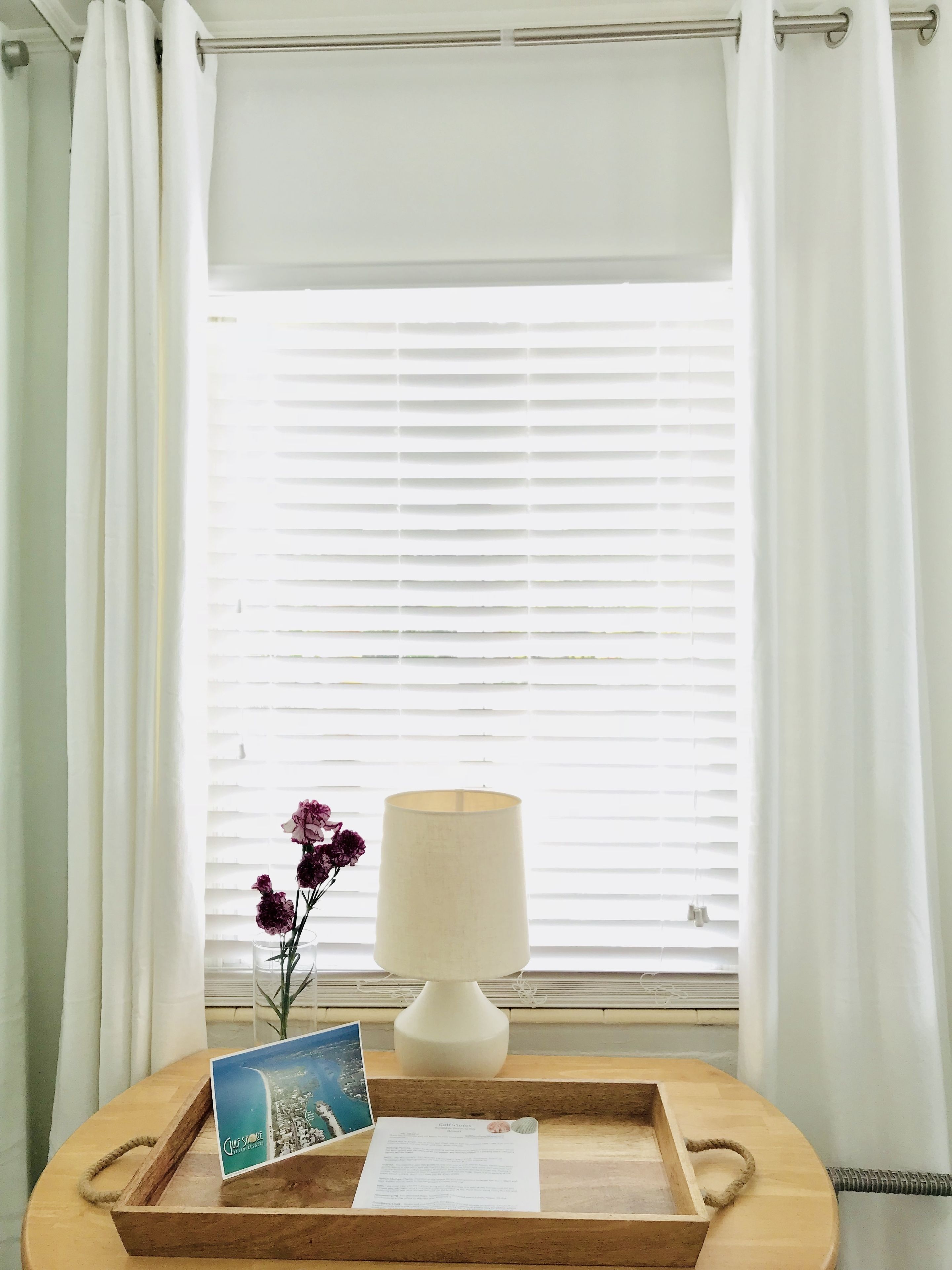 A wooden tray with a lamp, a postcard, and a menu on a table by a window with white curtains.