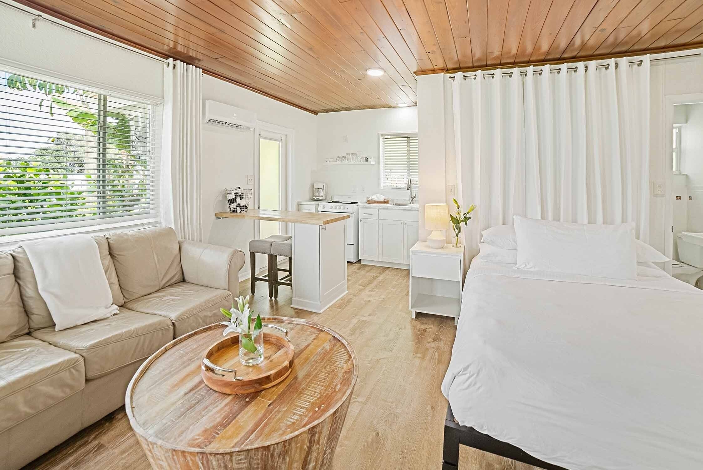 A modern hotel room with a wooden ceiling, a sofa, a bed, a dining area, and a kitchen.
