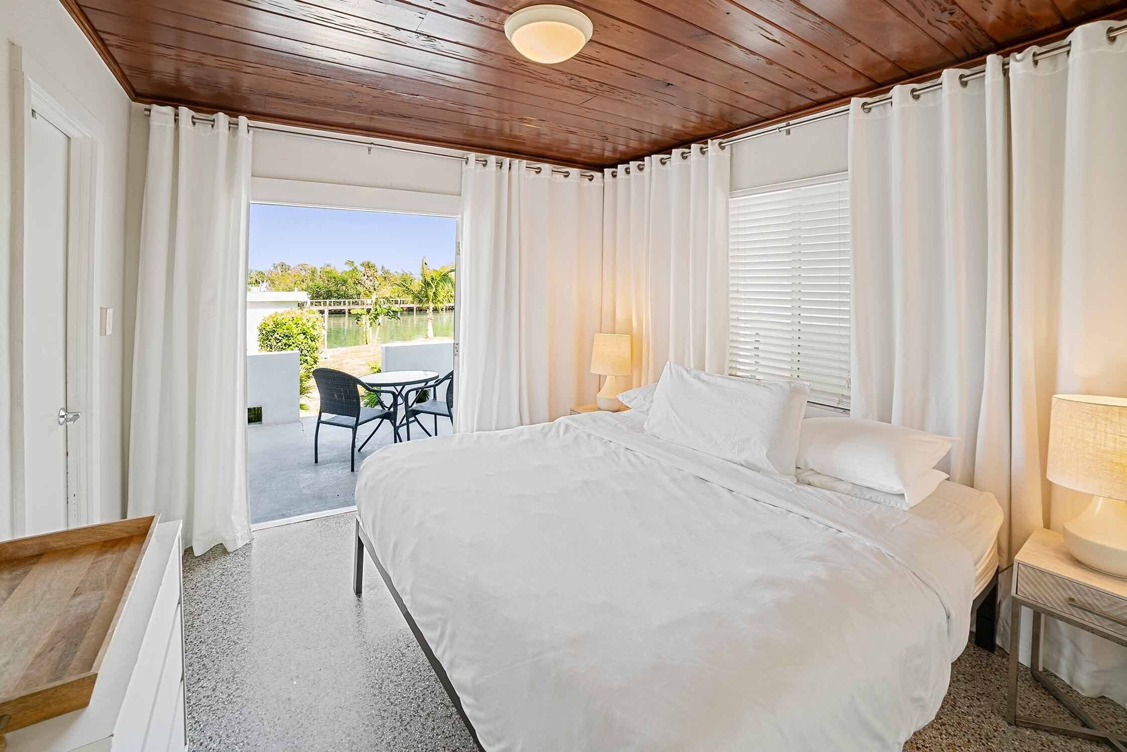 Quarto com uma cama grande, teto de madeira, cortinas brancas e vista para área externa com mesa e cadeiras.