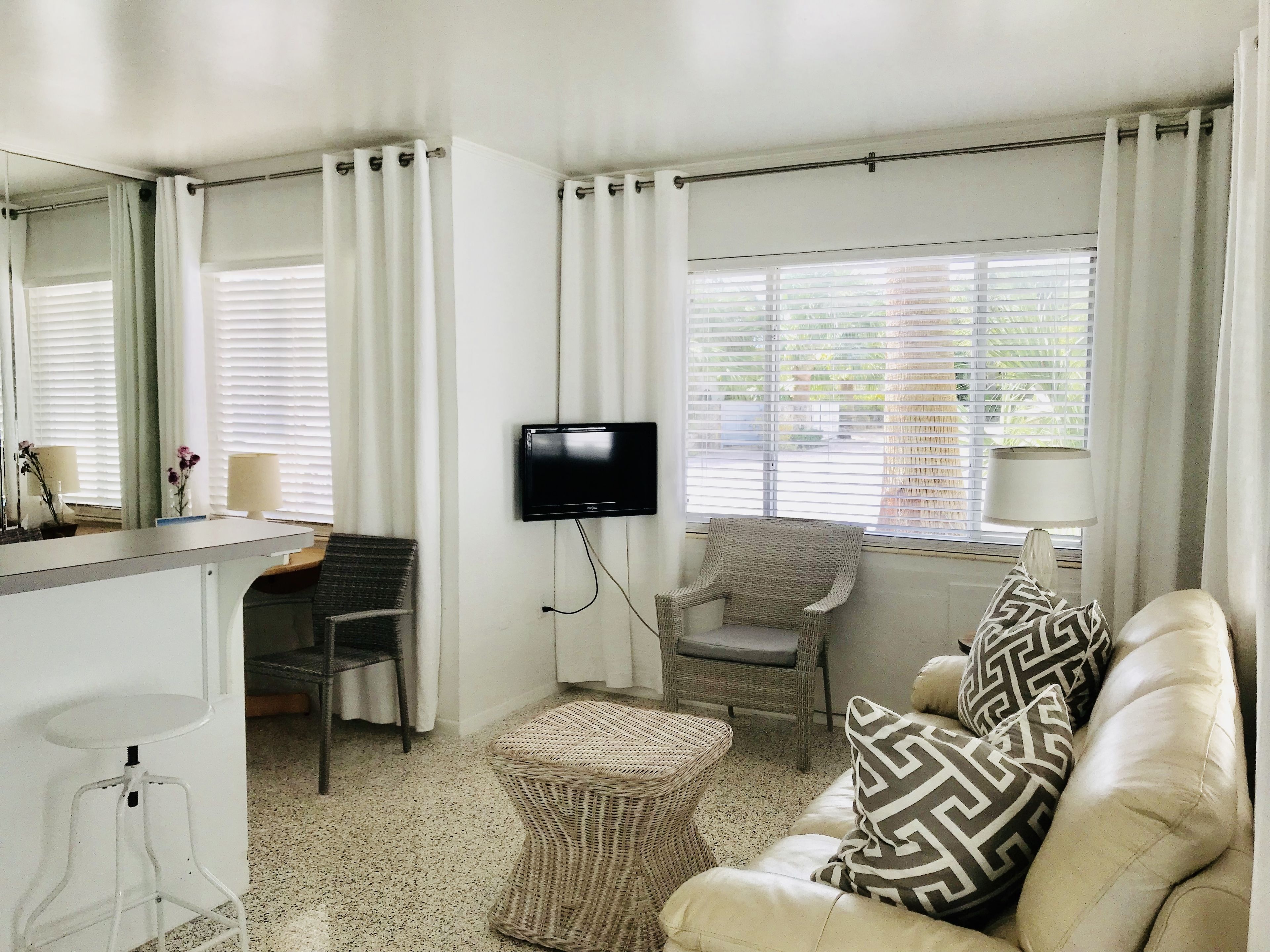 A room with a flat-screen TV mounted on the wall, a wicker chair, a sofa with patterned cushions, a small round table, and a bar area with a mirror and a lamp.