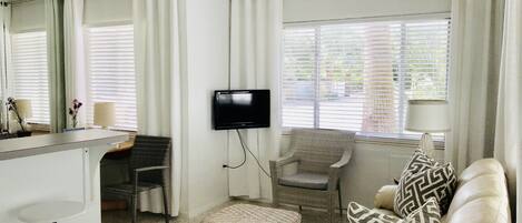 A room with a flat-screen TV mounted on the wall, a wicker chair, a sofa with patterned cushions, a small round table, and a bar area with a mirror and a lamp.