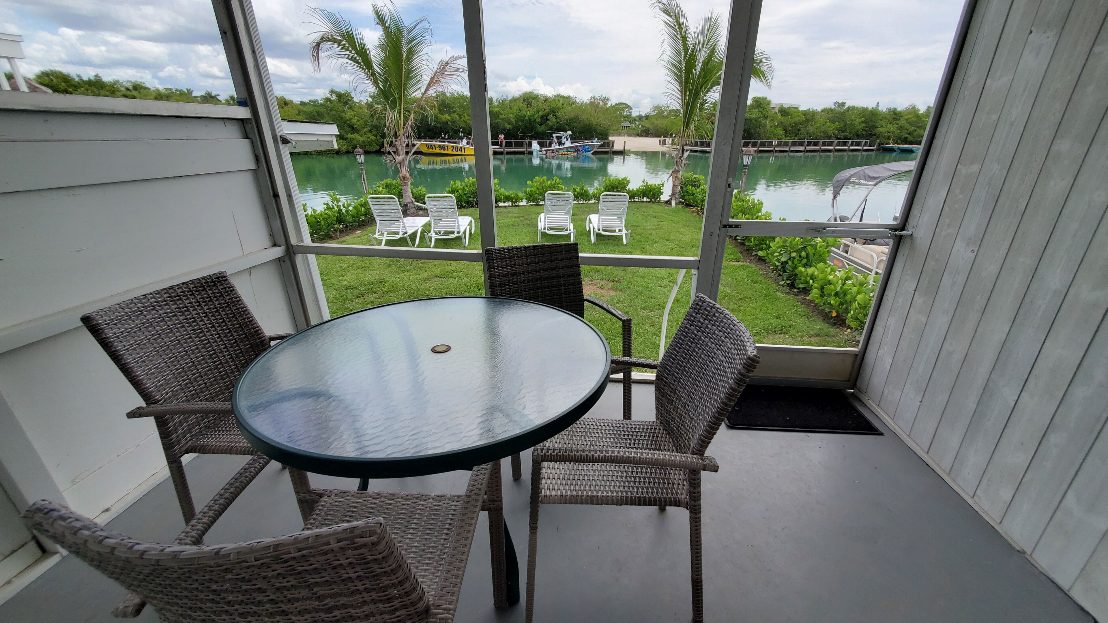 A balcony with wicker chairs and a glass table, overlooking a lake and a grassy area with chairs.