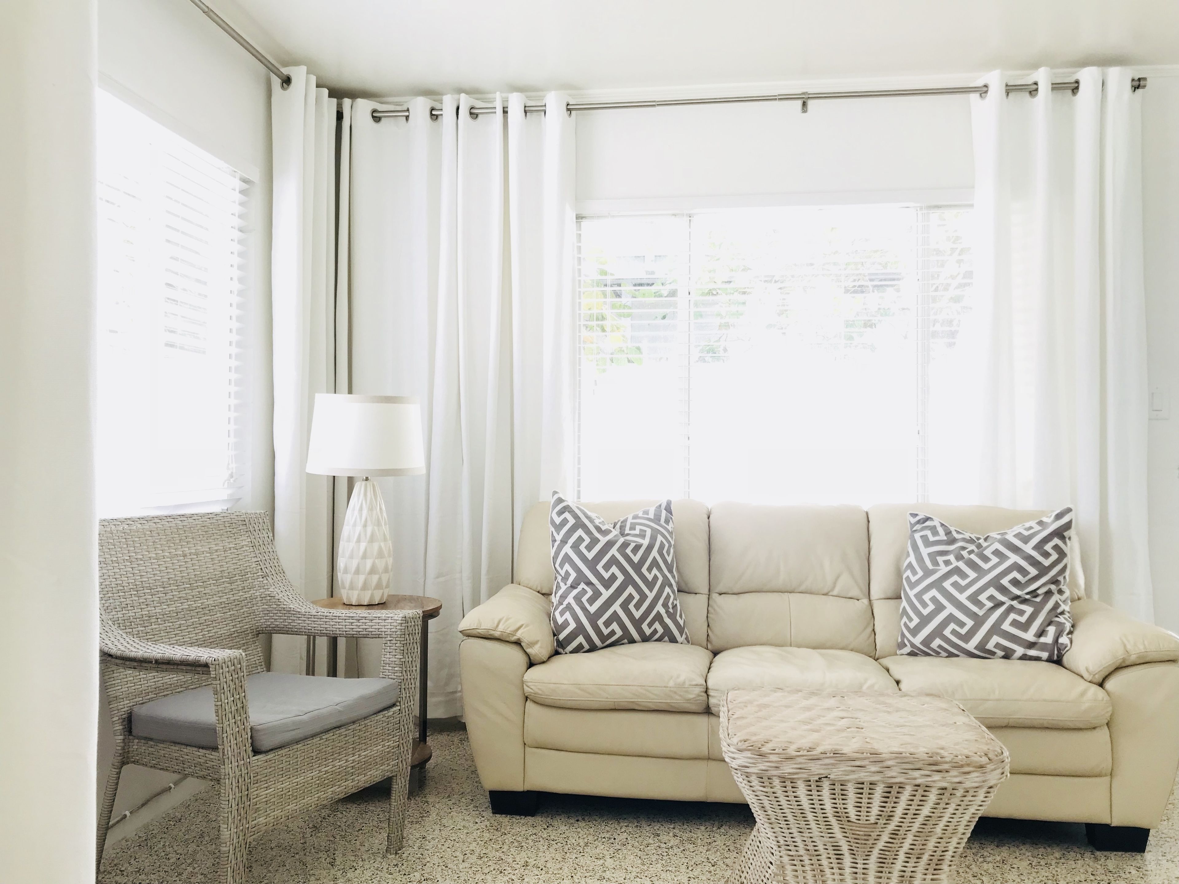 A living room with a beige sofa, two patterned pillows, a wicker chair, a small table with a lamp, and a window with white curtains.
