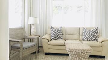 A living room with a beige sofa, two patterned pillows, a wicker chair, a small table with a lamp, and a window with white curtains.