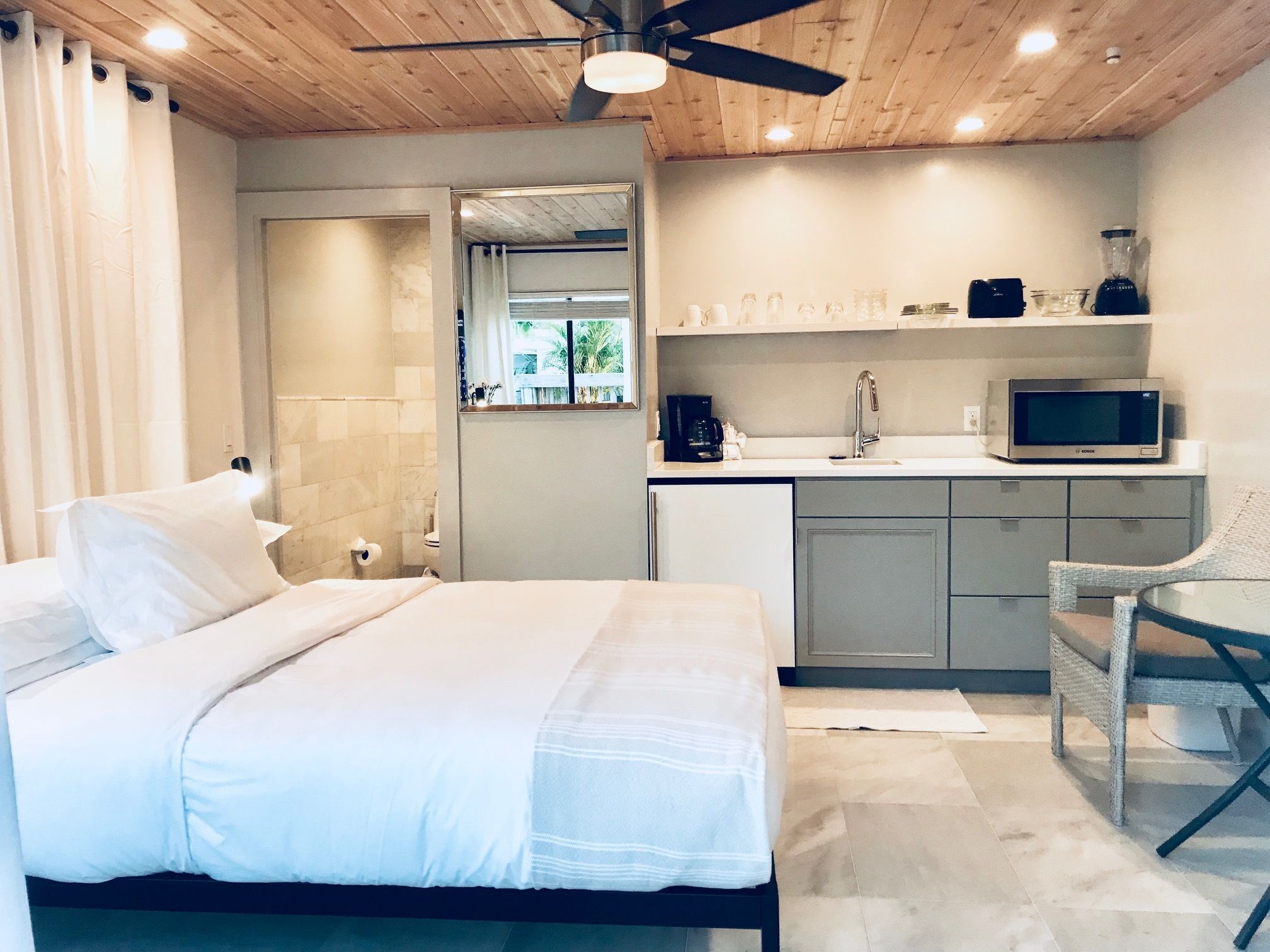 A bedroom with a bed, a chair, a kitchenette with a microwave, and a ceiling fan.