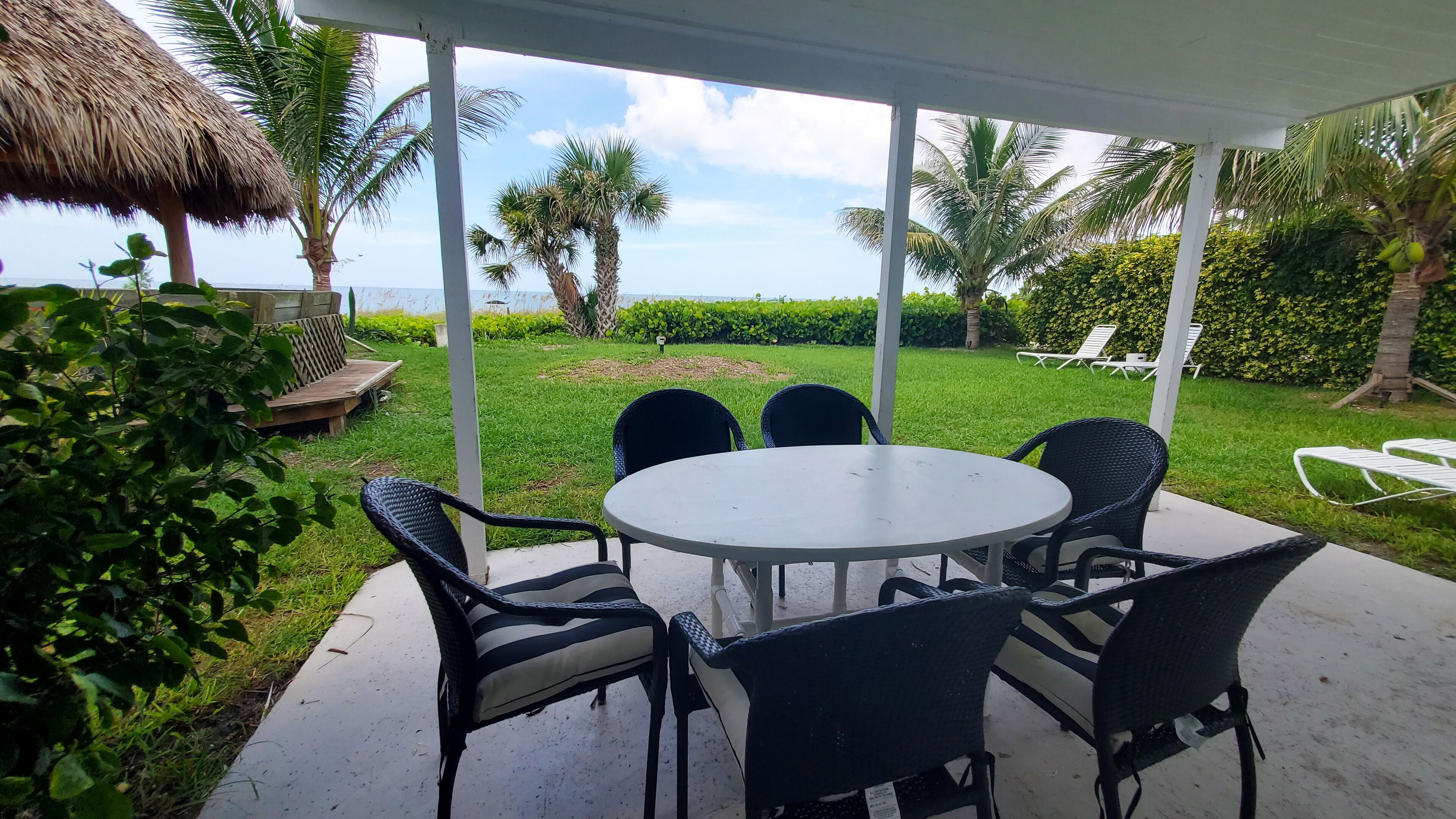 A covered outdoor patio with a round table and six chairs, surrounded by lush greenery and palm trees.
