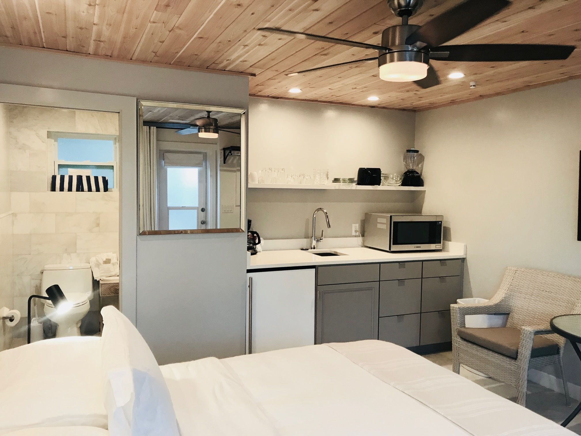 Habitación de hotel compacta con cama, kitchenette pequeña, zona de comedor con una silla y ventilador de techo.