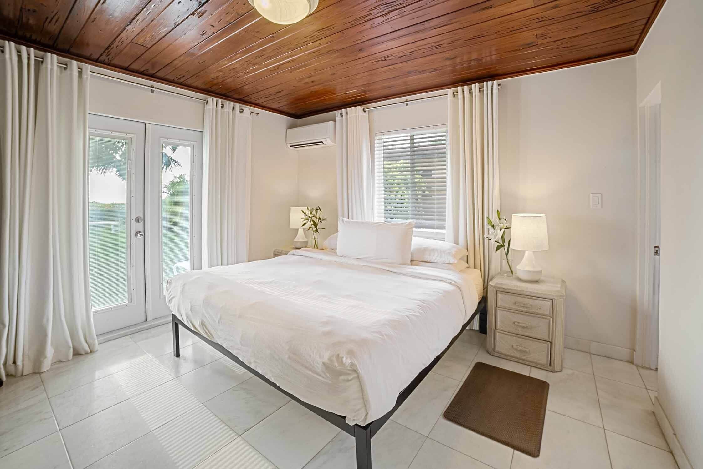 A bedroom with a wooden ceiling, a bed with white bedding, a nightstand with a lamp, and a window with blinds.